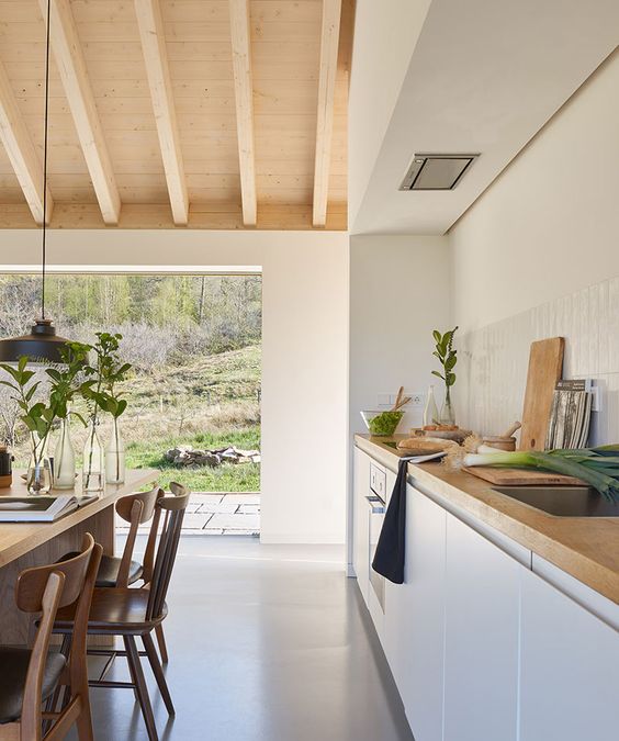a modern kitchen with white cabinetry, a dining zone with stained furniture, a pendant lamp and a wall that allows to open the space to outdoors