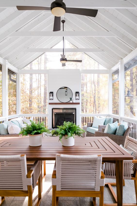 a modern screened patio with a fireplace, wicker furniture with light blue upholstery, a stained table and chairs and potted greenery