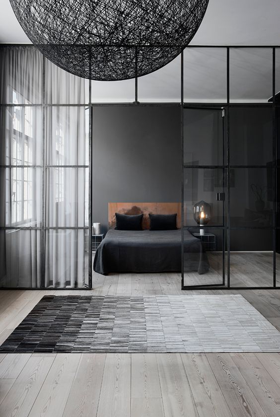 a moody Nordic bedroom with a black statement wall, a leather bed, nightstands and laps and a glass framed wall that separates it from the rest of the apartment