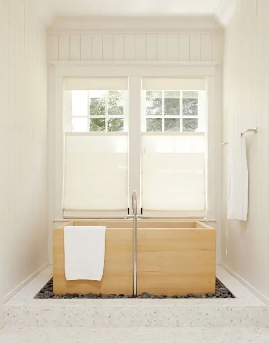 a neutral bathroom with a wood ofuro tub by the windows with shades placed into pebbles