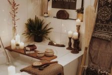 a neutral boho bathroom with large scale tiles, a tub clad with tiles, an artwork, lots of candles, some plants and lovely boho accessories