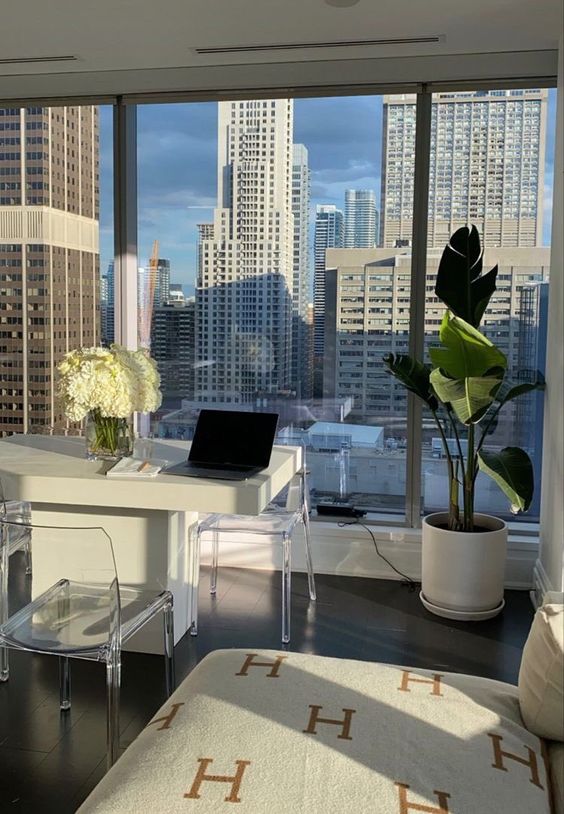 a pretty neutral home office with glazed wall and big city views, a white desk and actylic chairs, potted plants and blooms