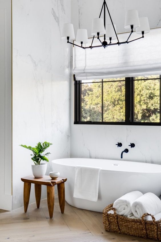 a refined modern neutral bathroom with a window, an oval tub, a wooden stool and a basket with towels and a chic chandelier