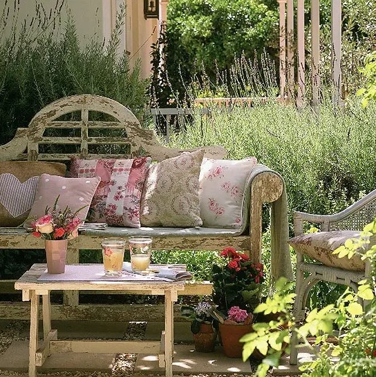 a refined shabby chic spring terrace with shabby chic furniture, potted plants and blooms and floral pillows and textiles and greenery around
