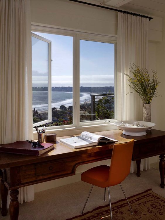 a romantic and dreamy home office with a window and coastal and sea views, a vintage stained desk, an orange chair and some books