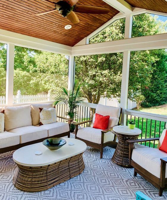 a screened patio with white seating furniture, coffee and side tables, bright pillows, greenery and a large printed rug