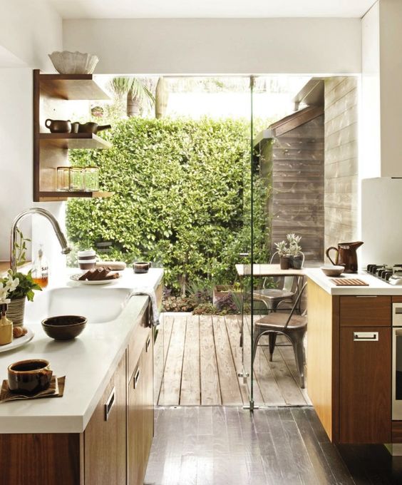a small modern kitchen with stained cabinets and a white stone countertop, open shelves and a glass wall that can open the space to a small deck