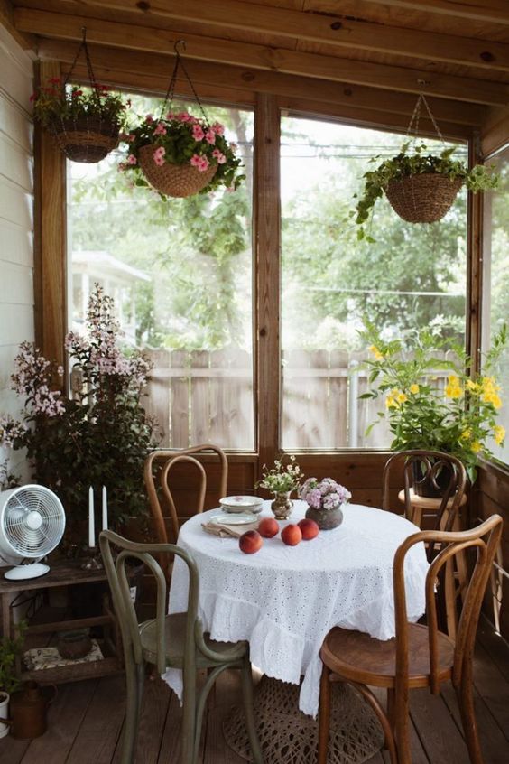 a small vintage screened porch with a round table and stained chairs, potted plants and greenery and a lot of light