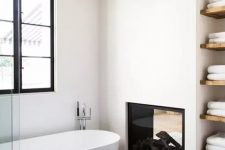 a stylish contemporary bathroom done in neutrals, with a double-sided fireplace, an oval tub, built-in shelves and towels