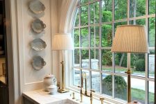 a vintage kitchen with light grey cabinets, white stone countertops, elegant table lamps and an arched window with a garden view