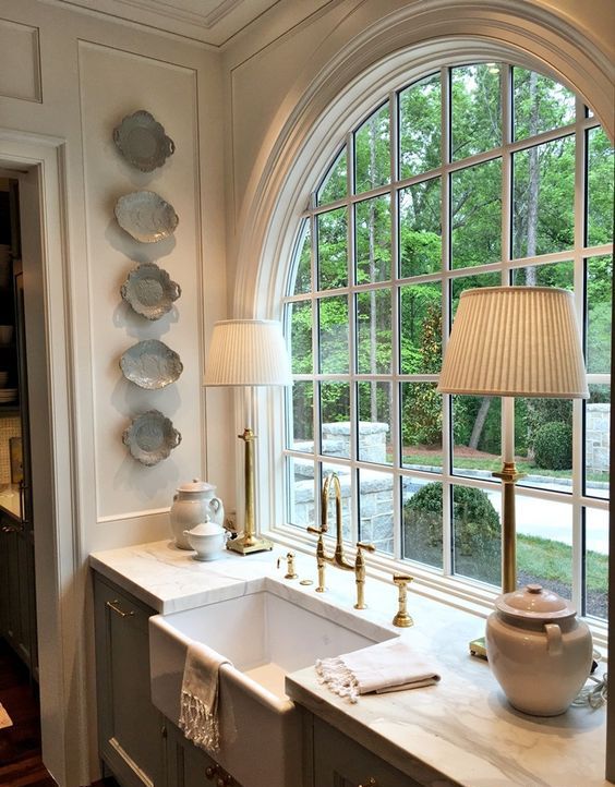 a vintage kitchen with light grey cabinets, white stone countertops, elegant table lamps and an arched window with a garden view