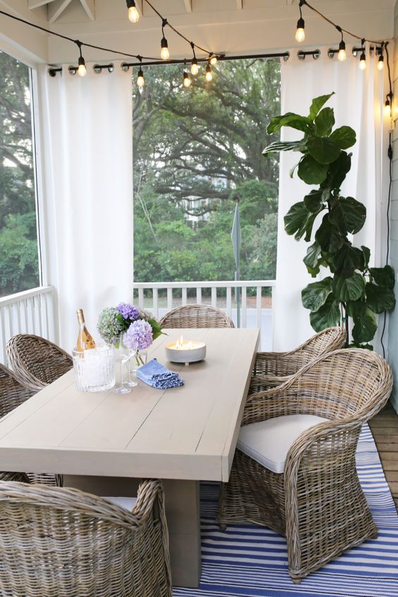 a welcoming screened porch with a beige wooden table, wicker chairs, string lights and potted plants
