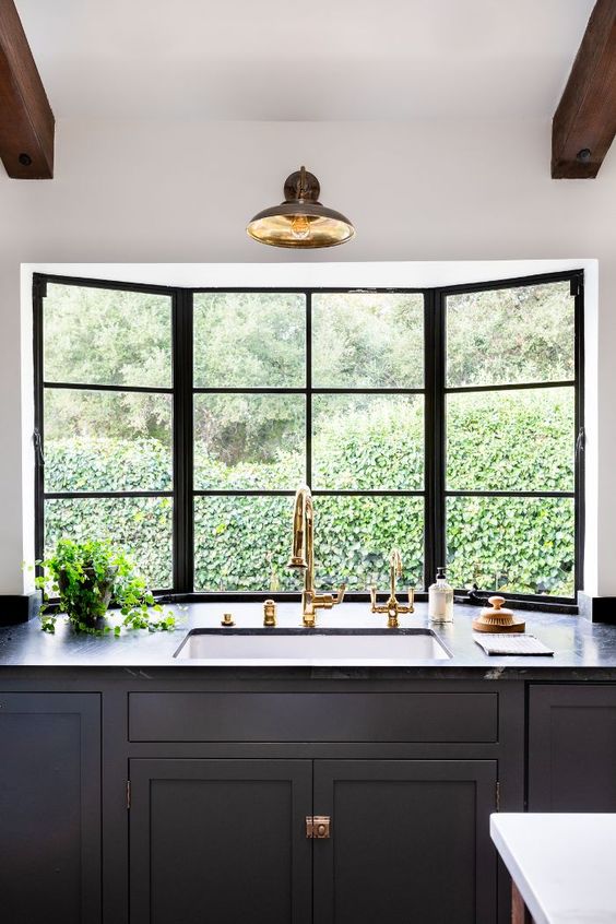 an elegant graphite grey kitchen with shaker style cabinets, grey marble countertops, brass touches and a bay window that opens on the garden