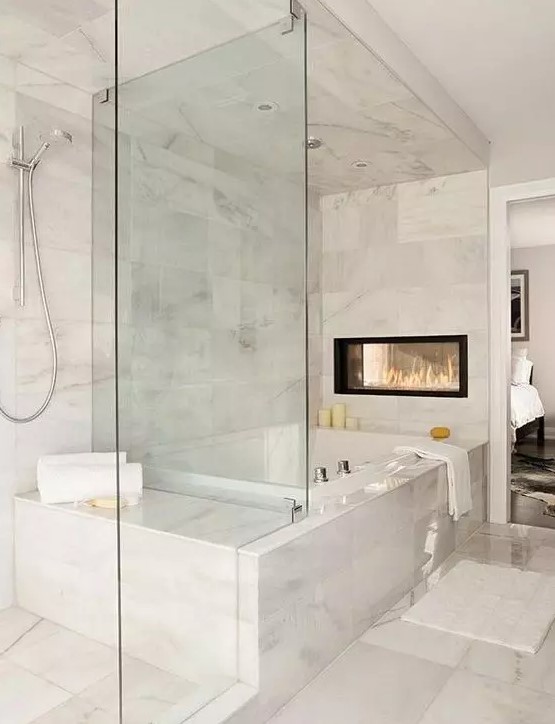 an exquisite white marble bathroom with a tub, a glass enclosed shower space, a double sided fireplace and some neutral textiles