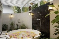 an outdoor bathtub of stone, with candle holders on the edge, with a shower, planted greenery and tall walls for privacy