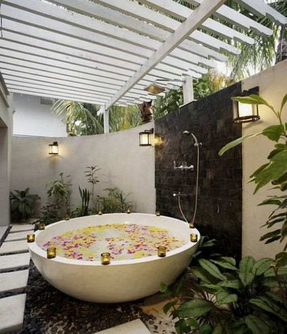 an outdoor bathtub of stone, with candle holders on the edge, with a shower, planted greenery and tall walls for privacy