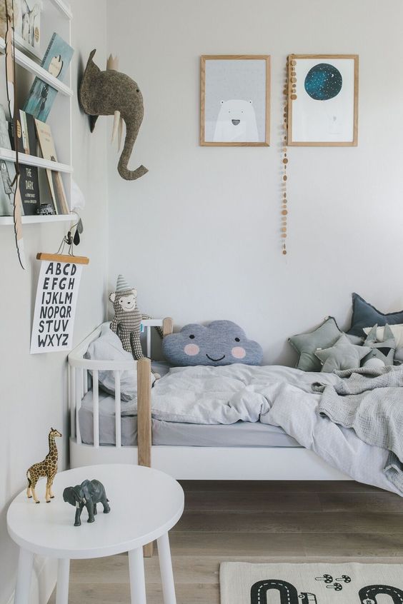 a Nordic kid's room with a white bed and a nightstand, ledges with books, pastel bedding and a gallery wall