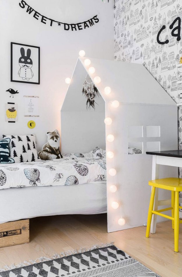 a Scandinavian kid's room with a bed with a house-shaped cover with lights, a desk, a yellow chair, a wall with printed wallpaper, some art