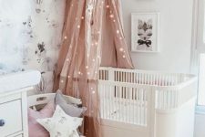 a Scandinavian kid’s room with a pastel wallpaper accent wall, a mini gallery wall, a white crib, a pink canopy with lights and star-shaped pillows
