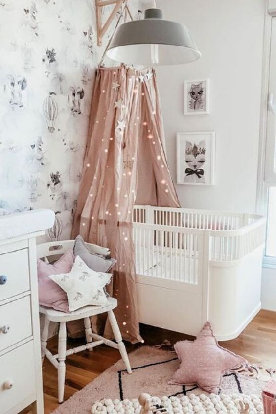 a Scandinavian kid's room with a pastel wallpaper accent wall, a mini gallery wall, a white crib, a pink canopy with lights and star-shaped pillows
