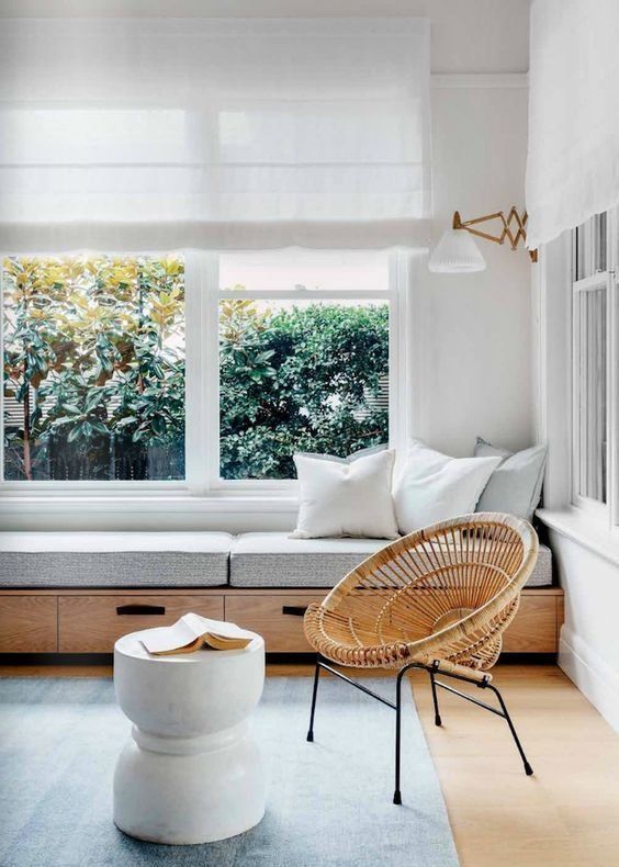 a beautiful contemporary space in light greys, with white Roman shades and a windowsill daybed with pillows, a rattan chair and a side table