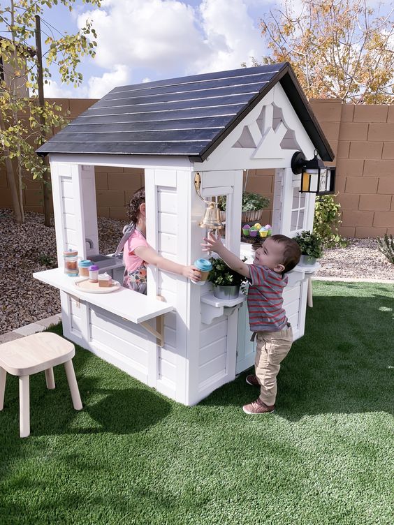 a black and white kids' playhouse with a black roof, with a shelf and a stool and potted greenery and blooms