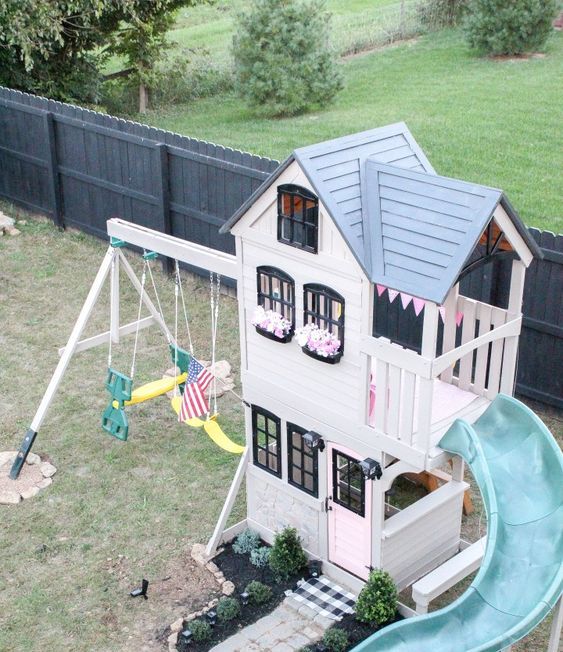 a black and white kids' playhouse with a grey roof, a turquoise slide, swings, greenery growing and a checked rug is all cool