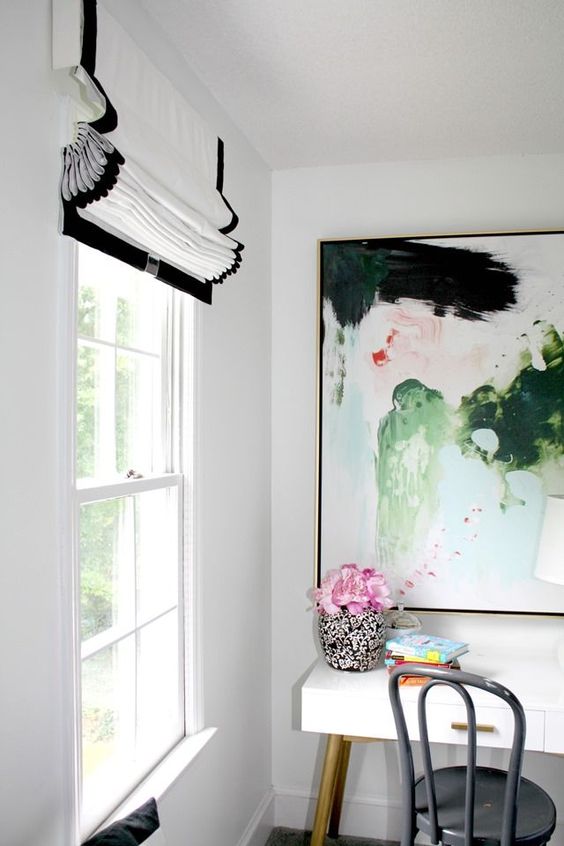 a bright home office nook with a white desk, a grey chair, a bold abstract artwork and a chic black and white Roman shade for a refined touch