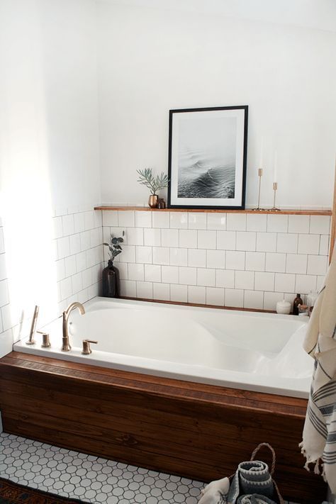 a chic neutral bathroom with white tiles of various sizes and shapes, a bathtub clad with rich-colored wood and a ledge with art and candles over the tub