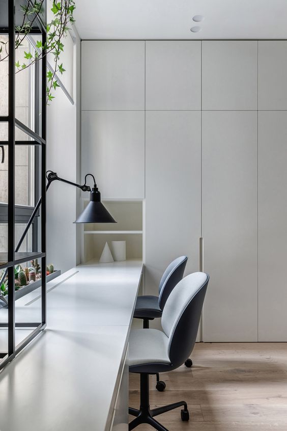 a clean minimalist home office with light grey storage units and a built-in shared desk, grey chairs and a table lamp is welcoming