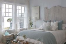 a coastal shabby chic bedroom with a wooden bed with a statement headboard, blue and white bedding, wooden furniture, a crystal chandelier and a wire bench