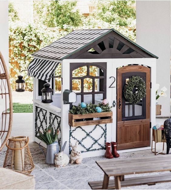 a cute outdoor kids' playhouse with a dark roof, with potted greenery and a wreath, some decor and lanterns is amazing