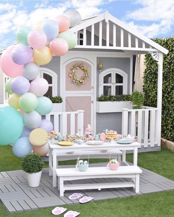 a grey and white kids' playhouse with potted greenery, a faux bloom wreath, an Easter tablescape and pastel balloons