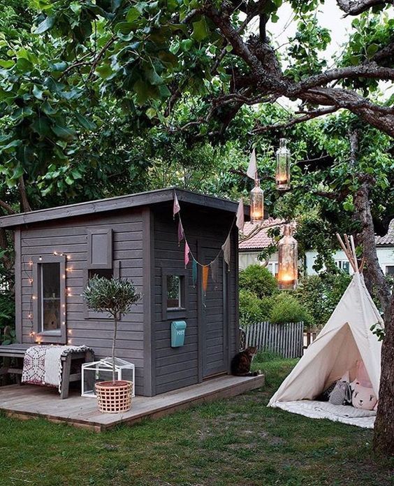 a grey playhouse with a colorful garland and lights, a potted tree and a teepee with pillows, hanging lamps