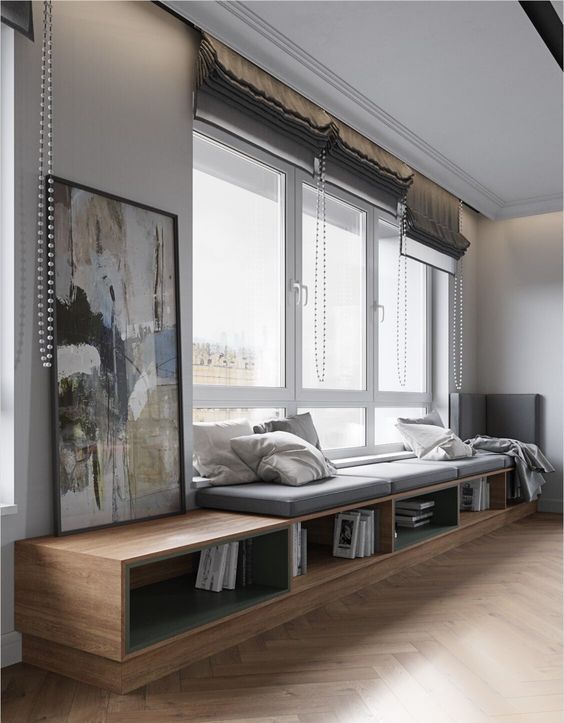 a large window with grey Roman shades and a large shelf with books that doubles as a daybed or a windowsill seat and styled with pillows