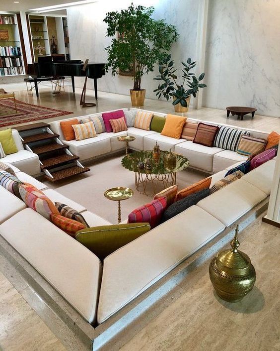 a lovely bold conversation pit with a sofa that lines it all, colorful pillows, a ladder and gold tables plus potted plants around