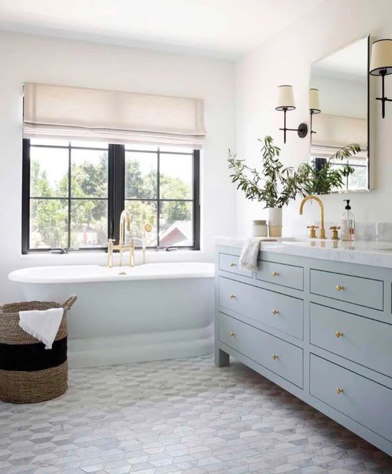 a lovely coastal bathroom with a hex floor, a light blue vanity, a mirror, gold touches and a neutral Roman shade to add interest to the space