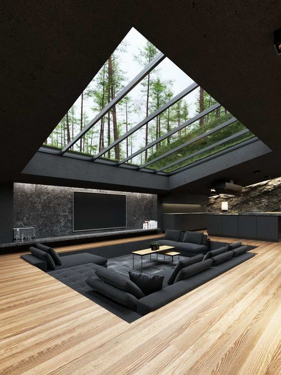 a minimalist conversation pit in black with a large sofa and pillows, a couple of coffee tables and a skylight through which you can see the trees