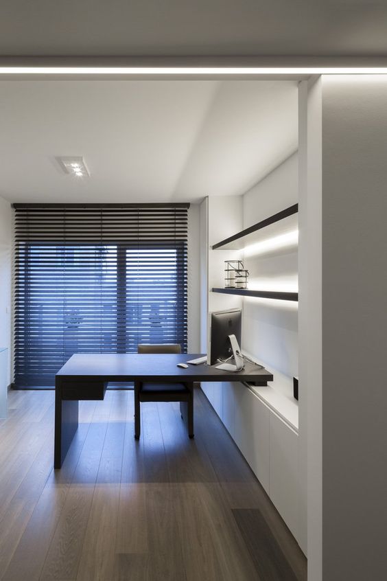 a minimalist home office with an open shelving unit with lights, a built-in desk and a blakc chair plus a floor to ceiling window