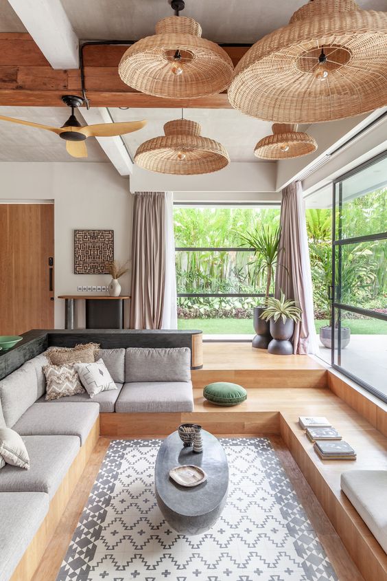 a modern meets boho conversation pit with a built-in bench with grey upholstery, an oval coffee table, woven pendant lamps