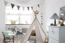 a neutral and pastel Nordic nursery with a teepee, a pastel blue dresser, a bed with printed bedding, a table and a chair, some lovely printed pillows