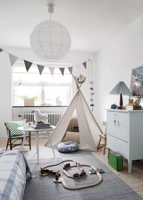 a neutral and pastel Nordic nursery with a teepee, a pastel blue dresser, a bed with printed bedding, a table and a chair, some lovely printed pillows