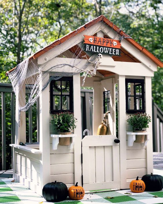 a neutral kids' playhouse with a roof, with black window frames, potted plants and blooms styled for Halloween