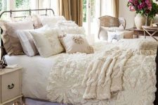 a neutral shabby chic bedroom with a metal bed, refined neutral furniture, ruffle and lace bedding and potted blooms