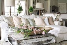 a neutral shabby chic living room with a white sectional, a low shabby table, greenery and blooms is welcoming