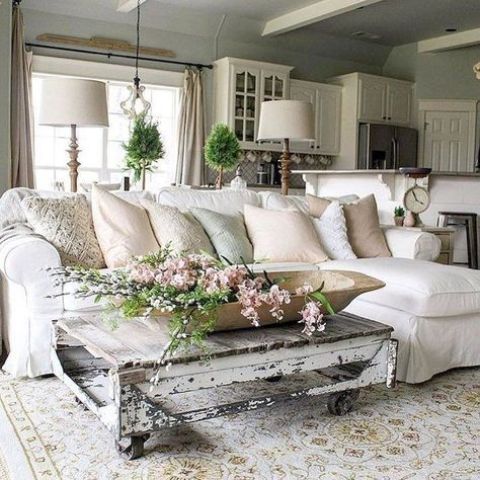a neutral shabby chic living room with a white sectional, a low shabby table, greenery and blooms is welcoming