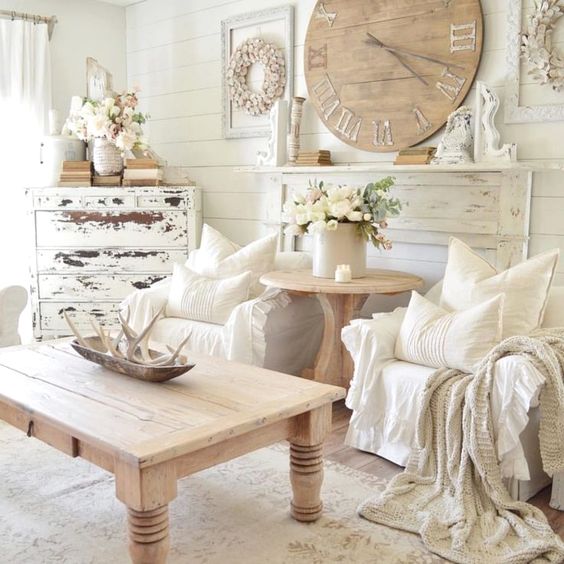 a neutral shabby chic living room with white walls, a shabby sideboard, a wooden clock and a table and some textiles