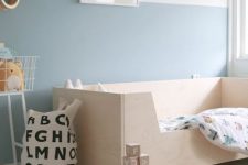 a pastel Scandinavian kid’s room with a plywood bed, color block blue walls, a colorful bunting, a printed fabric bag for storage and stylish toys
