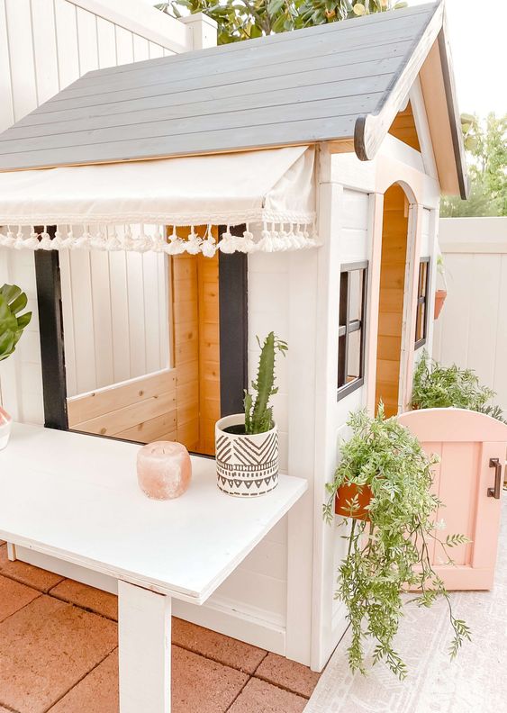 a pretty black and white kids' playhouse with a pink half door, with potted greenery and plants, with a large table next to it
