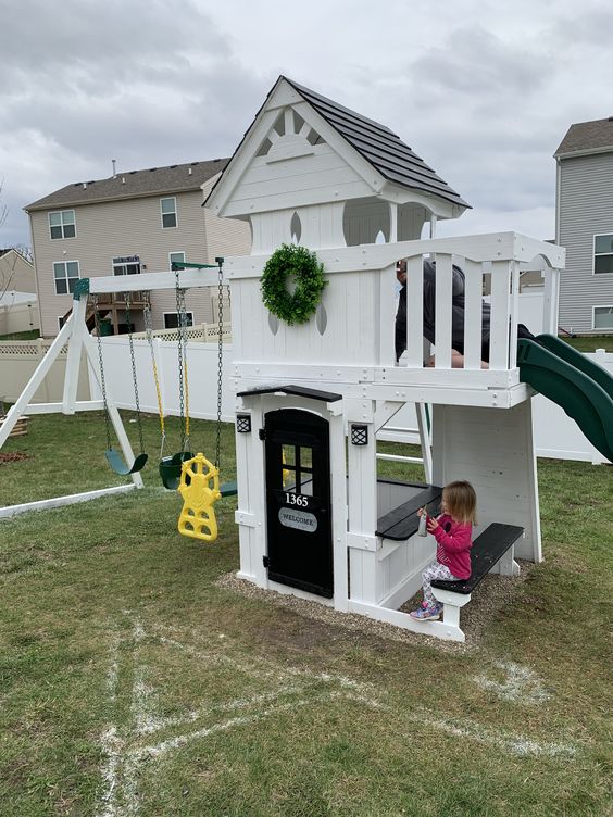 a pretty kids' playhouse of white planks, with a slide, a black door, a built-in dining space, swings and a greenery wreath